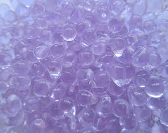 May include: A close-up of a pile of small, translucent, lavender-coloured glass beads. The beads are smooth and round, and they have a slight shimmer to them.