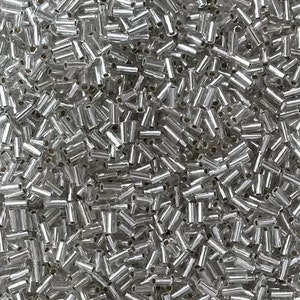 May include: A close-up shot of a pile of small, silver-colored, tube-shaped beads. The beads are densely packed together, creating a shimmering, reflective surface. The beads are uniform in size and shape, with a cylindrical form and a metallic sheen.