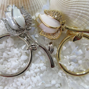 May include: Two decorative magnifying glasses with floral designs. One is silver-toned with a white flower and clear crystals, the other is gold-toned with a white and pink flower and clear crystals. The magnifying glasses are set against a white background.