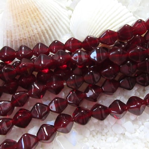 May include: Strands of dark red, faceted beads. The beads are diamond-shaped and strung together. Two white seashells are in the background, resting on a bed of white, granular material. The beads have a glossy finish.