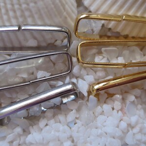 May include: Four metal belt loops, two silver and two gold, are arranged on a white background. The loops are rectangular and have a slight curve to them.