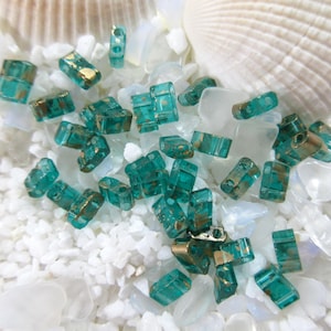 May include: A collection of teal and gold rectangular beads scattered on a bed of white crushed shells and glass. The beads have a metallic gold finish on the edges. A large, white seashell is in the background.