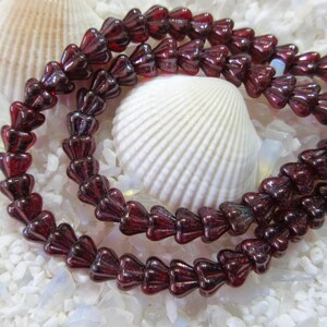 May include: Strands of dark red glass beads with a flower-like shape. The beads are arranged in rows, with a glossy finish. The background features white seashells and small white stones, creating a beachy aesthetic. Ideal for jewelry making.