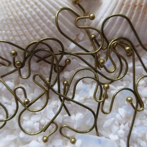 May include: A close-up of a pile of gold-toned earring hooks. The hooks are made of metal and have a small ball at the end. The hooks are scattered on a white surface.