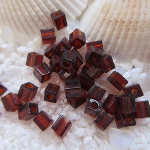 May include: A close-up of a pile of small, square, dark brown glass beads. The beads are scattered on a white, grainy surface.