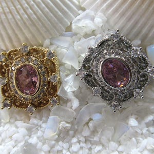 May include: Two ornate rings with pink gemstones set in a silver and gold filigree design. The rings are displayed on a bed of white pebbles.