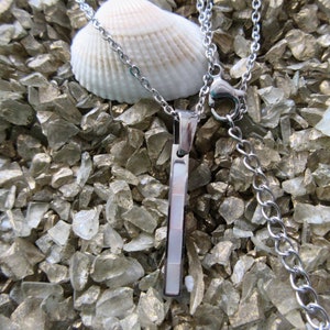 May include: A silver necklace with a rectangular pendant featuring a white mother-of-pearl inlay. The necklace is displayed on a bed of white and silver glitter.