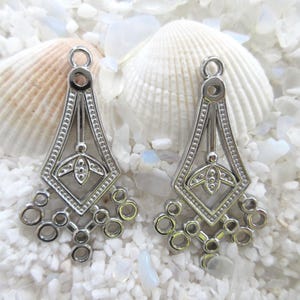 May include: Two silver-colored chandelier earring components with an Art Deco design. Each component features a geometric shape with a floral motif and a series of small circles at the bottom. The earrings are displayed on a bed of white pebbles and seashells.