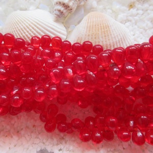 May include: A strand of bright red glass beads, each bead is shaped like a teardrop. The beads are arranged in a loose pile on a white surface.