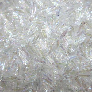 May include: Close-up of a pile of clear, iridescent glass beads. The beads are small, rectangular, and have a slightly irregular shape. They reflect light, creating a shimmering effect. These beads are suitable for jewelry making and craft projects.