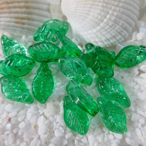 May include: A collection of translucent green glass leaf beads. The beads are scattered on a bed of white pebbles, with a white seashell in the background. The leaves have detailed veining.