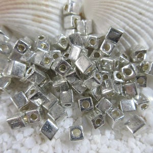May include: A collection of small, silver-colored cube beads with a hole in the center of each face. The beads are scattered on a bed of white, granular material, with two seashells in the background.