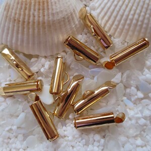May include: A group of ten gold-colored metal cord end caps, scattered on a white background. The caps are cylindrical with a small opening at one end.