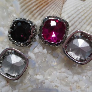 May include: Four square-shaped, silver-toned, decorative embellishments with a scalloped edge. The embellishments have clear, pink, and black faceted glass stones.