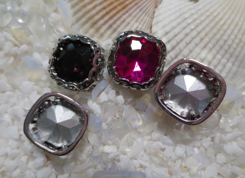 May include: Four square-shaped, silver-toned, decorative embellishments with a scalloped edge. The embellishments have clear, pink, and black faceted glass stones.