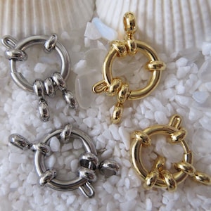 May include: Four silver and gold metal clasps with a round ring and a decorative knot design. The clasps are photographed on a white background.