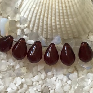 May include: Seven dark red glass teardrop beads strung on a silver wire. The beads are arranged in a row and are all the same size and shape.