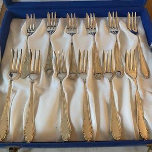 May include: A set of twelve silver-toned metal forks with four tines and decorative handles, arranged in two rows within a blue presentation box lined with white fabric. Each fork features a slender neck and a flared, detailed handle base.