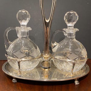 May include: A silver-plated cruet set featuring two clear glass bottles with faceted stoppers and handles. The bottles sit in a metal frame with a central carrying handle, all resting on an oval silver-plated tray with small decorative feet.