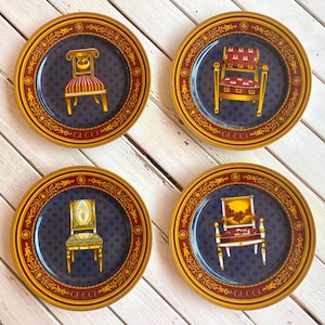 May include: Four decorative plates with a navy blue background and gold trim. Each plate features a different chair design in the center. The plates have a gold border with the word "GUCCI" in red.