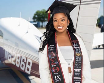 May include: A graduate in a cap and gown poses in front of a small airplane. She wears a white blazer and a sash with text including "USC BA Class of 2025" and "21 SAVAGE". The background is a clear blue sky.