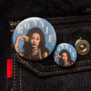 May include: Two round blue badges with the name "RONNIE" in white letters. Each badge features a portrait of a woman with dark hair. The badges are on a dark blue denim background.