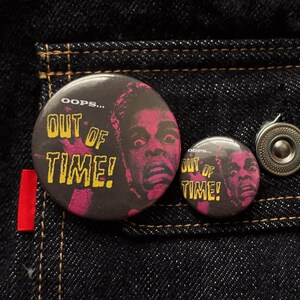May include: Two black and pink round buttons with the text "OOPS... OUT OF TIME!" and a graphic of a person's face. The buttons are on a dark blue denim background with a red tag and a silver button.