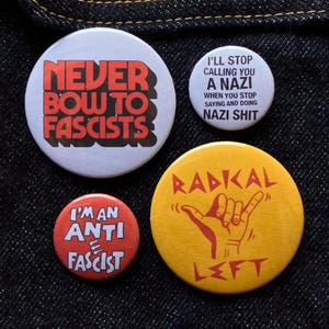 May include: Four round buttons with anti-fascist messages. One button says "NEVER BOW TO FASCISTS" in red on white. Another says "I'M AN ANTI FASCIST" in white on red. A third says "I'LL STOP CALLING YOU A NAZI" on white. The last button is yellow with a hand gesture and the word "RADICAL LEFT".
