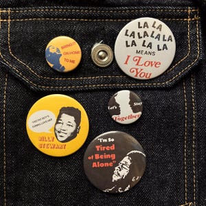 May include: A selection of circular pin badges with diverse designs and text, including "Bring It On Home To Me," "La La La Means I Love You," and a portrait of Billy Stewart. The badges are displayed on a dark blue denim fabric.