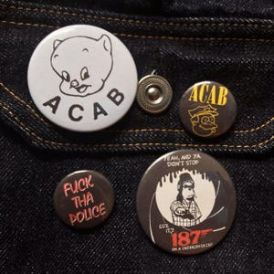 May include: A collection of five round buttons with various designs and text. One button features a cartoon pig with the letters "ACAB". Another says "FUCK THA POLICE". The buttons are on a dark blue denim background.