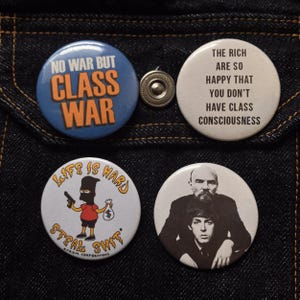 May include: Four circular pin badges with different designs. One badge has the text "NO WAR BUT CLASS WAR" in blue and orange. Another says "THE RICH ARE SO HAPPY THAT YOU DON'T HAVE CLASS CONSCIOUSNESS". A third features a cartoon character and the text "LIFE IS HARD STEAL SHIT". The last badge has a black and white image of two men.