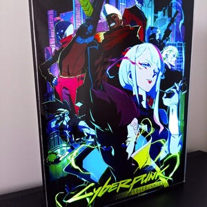 May include: A rectangular light box featuring a vibrant Cyberpunk Edgerunners graphic with multiple characters in a neon-lit city. The artwork displays a dark colour palette with bright yellow, blue, and magenta accents, and the title text Cyberpunk Edgerunners is printed in yellow at the bottom.