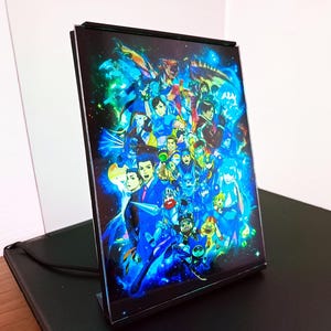 May include: A rectangular acrylic panel featuring a vibrant collage of anime characters in shades of blue, green, and yellow. The artwork is set against a cosmic background and is displayed on a black stand.