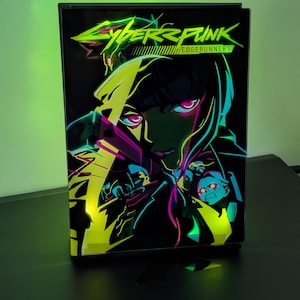 May include: A rectangular light box featuring the Cyberpunk Edgerunners logo in neon green at the top. The graphic displays stylised anime characters with glowing pink eyes and vibrant yellow and teal accents against a deep black background.