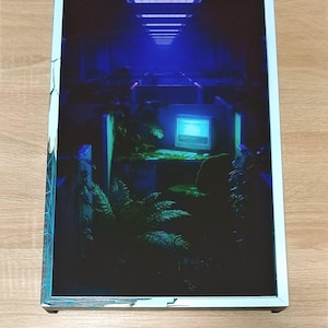 May include: A framed print showcasing a retro-futuristic office scene in blue tones. The image includes a computer, plants, and a hallway with overhead lighting. The white frame has a teal border. The print is displayed on a wooden surface.