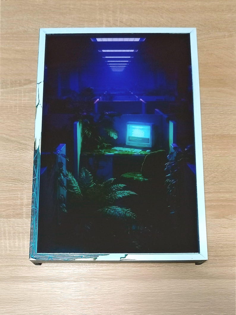 May include: A framed print showcasing a retro-futuristic office scene in blue tones. The image includes a computer, plants, and a hallway with overhead lighting. The white frame has a teal border. The print is displayed on a wooden surface.