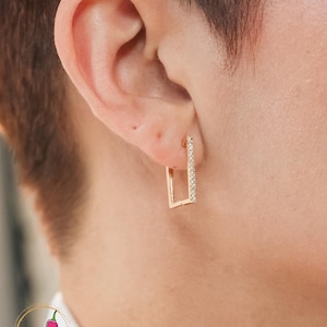 May include: Close-up of a gold rectangular earring with a row of small, sparkling stones along one side. The earring is worn on the ear of a person with short, dark hair. The earring is a simple, elegant design.