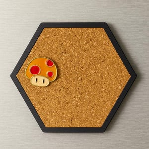 May include: Hexagon-shaped corkboard with a black frame. A colorful mushroom pin with red spots and a cream-colored base is attached to the corkboard. The corkboard is a light brown color.
