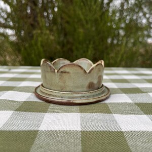 Pode incluir: Uma pequena taça de cerâmica redonda com uma borda recortada em forma de pétalas. A peça apresenta um esmalte salpicado em verde sálvia claro com subtis detalhes castanhos ao longo das bordas e da base. Assenta num pires de cerâmica a combinar.