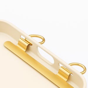 May include: A cream-colored tray with gold-colored accents. The tray features a long, rectangular gold bar and two gold ring handles. The design is minimalist and modern, with a focus on clean lines and a neutral color palette.