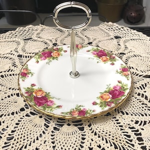 May include: A two-tiered serving tray with a silver handle. The white porcelain plates are decorated with a floral pattern of pink and yellow roses and a gold rim. The tray is set on a cream-colored, crocheted doily.
