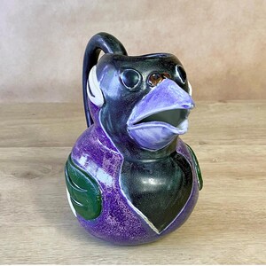 May include: A whimsical ceramic pitcher shaped like a bird, featuring a black head and body with purple and green accents. The pitcher has a curved handle and an open beak, perfect for serving beverages.