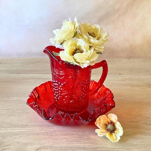 May include: A vintage red glass pitcher and matching bowl set. The pitcher has a handle and is filled with yellow flowers. The bowl has a ruffled edge. The set is on a wooden surface.