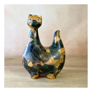 May include: A ceramic cat figurine with a long neck and a tail. The cat is decorated with a patchwork pattern of dark blue, green, and tan shapes. The cat's head is tan with black and white details.