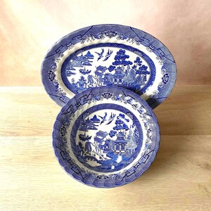 May include: Two blue and white ceramic dishes. The oval platter and round bowl feature a detailed landscape design with pagodas, birds, and water elements. The rims have a decorative border. The dishes are placed on a light wood surface.