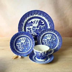 May include: A blue and white Willow pattern china set, including plates, bowls, a teacup, and saucer. The set features a classic landscape design with pagodas, bridges, and birds. The china is arranged on a wooden surface.