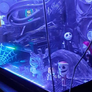 May include: A custom-built computer tower with a clear glass panel, illuminated by blue LED lights. Inside, there are fans, a graphics card, and figurines from The Nightmare Before Christmas. A spiderweb graphic is visible.