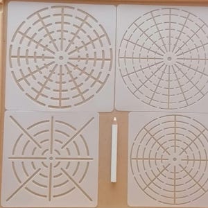 Op de afbeelding: Vier witte sjablonen met cirkelvormige geometrische ontwerpen en een witte potlood. De sjabels zijn gerangschikt in een vierkante formatie in een houten bak. De ontwerpen bevatten concentrische cirkels en radiale lijnen.
