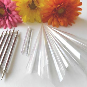 Puede incluir: Un surtido de materiales de manualidades, que incluyen herramientas de punteado de doble punta con purpurina roja, varitas de purpurina y láminas transparentes en forma de cono. Margaritas gerbera rosas, amarillas y naranjas brillantes en la parte superior.