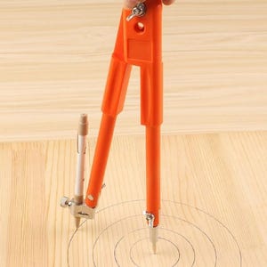 May include: An orange compass with a pencil attached is used to draw concentric circles on a light wood surface. The compass is held by a hand, and the circles are drawn in black ink. The compass is a drafting tool.